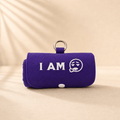 A purple rolled-up fabric pouch or case with a metal ring handle, featuring the white embroidered text 'I AM' and a sad face graphic, set against a warm beige background with soft leaf shadows. This product called Burritote.