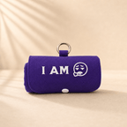 A purple rolled-up fabric pouch or case with a metal ring handle, featuring the white embroidered text 'I AM' and a sad face graphic, set against a warm beige background with soft leaf shadows. This product called Burritote.