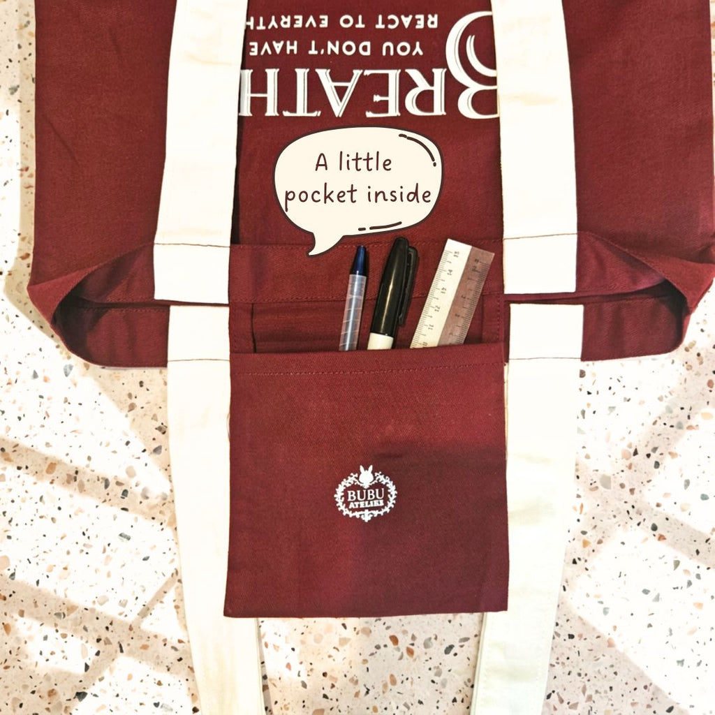 A maroon canvas tote bag with white colored straps and a front utility pocket. The pocket contains pens, a ruler, and a small notebook, and features a white bunny logo with the text 'BUBU Atelier'.