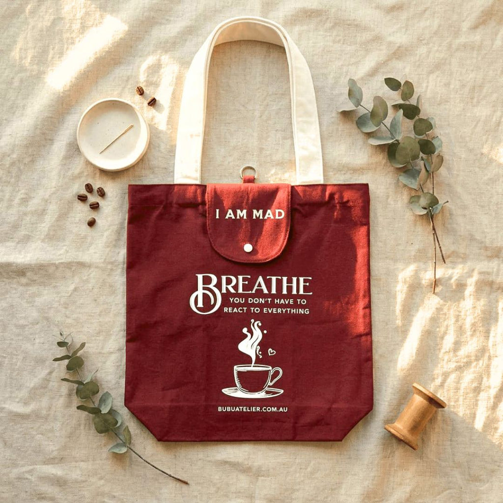A burgundy tote bag with white handles and printed white text reading 'I AM MAD BREATHE YOU DON'T HAVE TO REACT TO EVERYTHING' with a coffee cup graphic, styled on a beige fabric surface with eucalyptus leaves, a small dish, coffee beans, and a wooden spool.