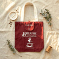 A burgundy tote bag with white handles and printed white text reading 'I AM MAD BREATHE YOU DON'T HAVE TO REACT TO EVERYTHING' with a coffee cup graphic, styled on a beige fabric surface with eucalyptus leaves, a small dish, coffee beans, and a wooden spool.