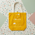 A bright yellow canvas tote bag with white handles and printed text reading 'I AM PAUSE' and a simple line drawing of a sleeping figure, photographed from above on a terrazzo-patterned surface with pastel color blocks. Made by Bubu Atelier.