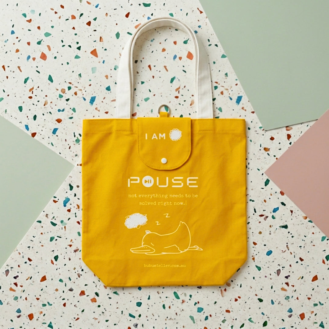 A bright yellow canvas tote bag with white handles and printed text reading 'I AM PAUSE' and a simple line drawing of a sleeping figure, photographed from above on a terrazzo-patterned surface with pastel color blocks. Made by Bubu Atelier.
