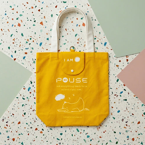 A bright yellow canvas tote bag with white handles and printed text reading 'I AM PAUSE' and a simple line drawing of a sleeping figure, photographed from above on a terrazzo-patterned surface with pastel color blocks. Made by Bubu Atelier.