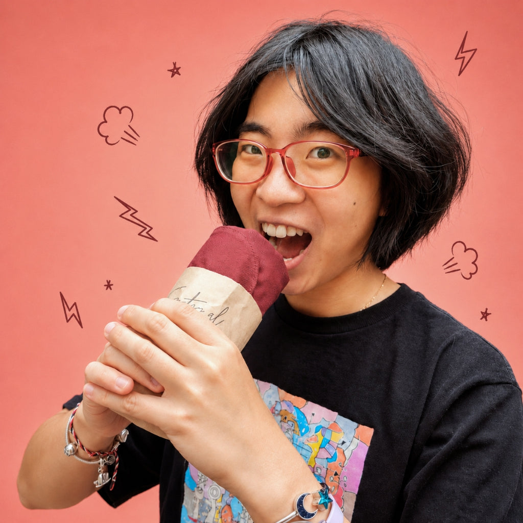 A person with short black hair and red-framed glasses holds a paper-wrapped drink or snack against a coral-pink background with simple doodle graphics.