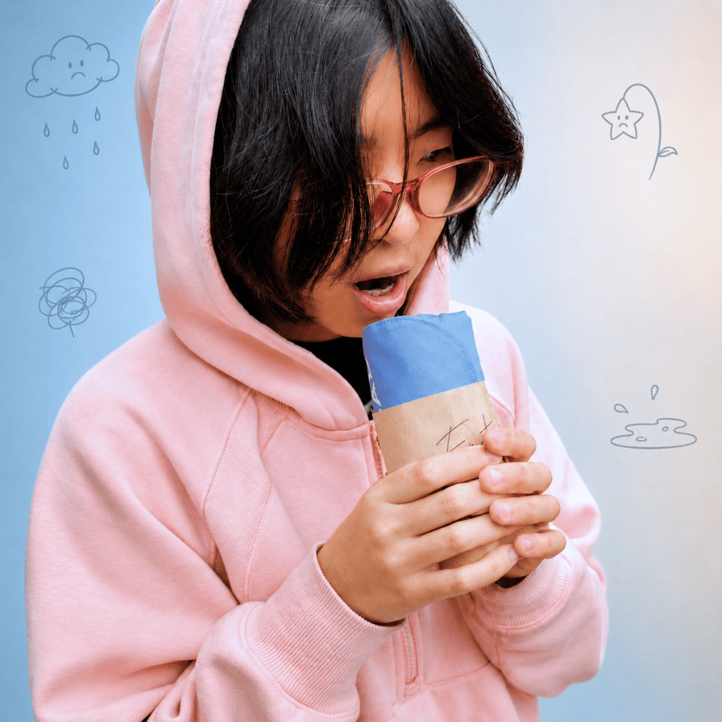 A person wearing a light pink hoodie and glasses holds a cup wrapped in blue and beige paper against a light blue background with simple doodled illustrations.