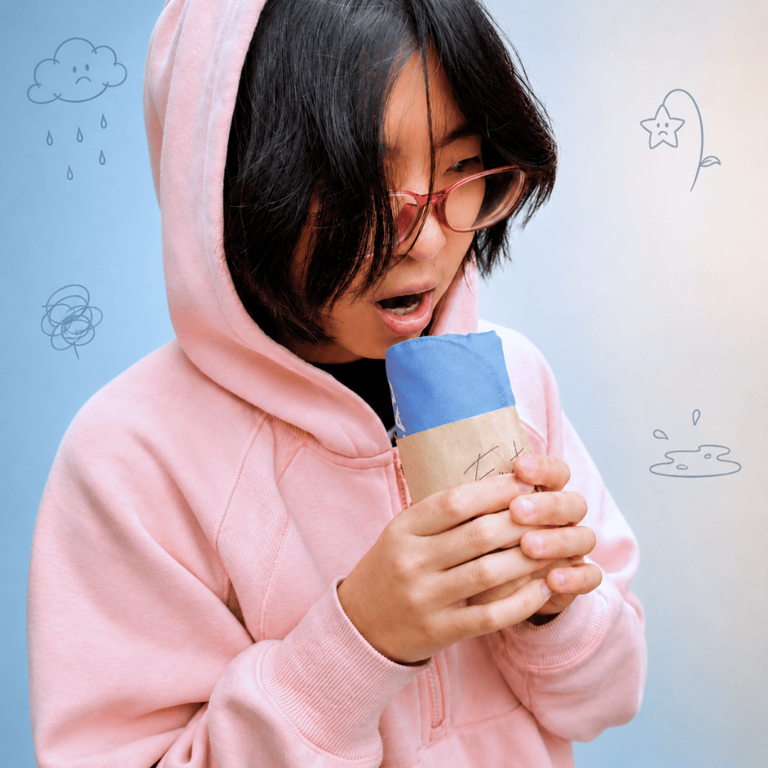 A person wearing a light pink hoodie and glasses holds a cup wrapped in blue and beige paper against a light blue background with simple doodled illustrations.
